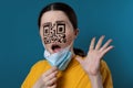 Portrait of a shocked woman with a QR code instead of eyes and nose, removing protective mask from her face. Blue