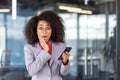 Portrait of shocked young African American woman in business suit standing in office, holding mobile phone, looking at Royalty Free Stock Photo