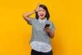 Portrait of shocked young Asian woman hand on head with disappointed expression and holding smartphone on yellow background