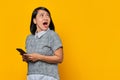 Portrait of shocked young Asian woman holding mobile phone and looking aside on yellow background Royalty Free Stock Photo