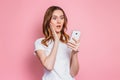 Portrait of a shocked young girl looking at phone and reads a messages or news  over pink background. Surprised woman in a Royalty Free Stock Photo