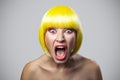 Portrait of shocked young woman with freckles, red makeup and yellow wig looking at camera with unbelievable face and screaming