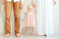 Portrait of shoeless mother and father holding hands of smiling baby girl in pink holiday dress on white background