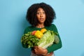 Portrait shot of amazed brunette young woman with bushy hair bites lips and carries cluster of different fresh veggies