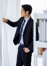 Portrait shot of Asian young company bookkeeper in formal black suit and necktie standing hold hand open clear white curtain look