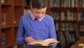 Portrait shot of the cute schoolboy and standing near the bookshelf in the library and reading a book. Royalty Free Stock Photo