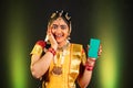 Portrait shot of Excited happy Bharatnatyam dancer showing green screen mobile phone by looking at camera on stage -