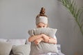 Portrait of shy cute little girl with bun hairstyle sitting in bedroom on bed with pillow hiding her face wearing sleeping mask Royalty Free Stock Photo