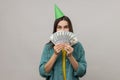 Shy woman with party cone on head covering half of face with money, lottery win, big profit. Royalty Free Stock Photo