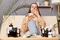 Portrait of sick sleepy woman wearing sweater and wrapped in scarf sitting on sofa at home, drinking hot tea from white cup,