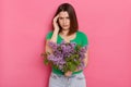 Portrait of sick young girl with brown hair wearing green casual t shirt holding bouquet of lilac flowers, suffering headache,