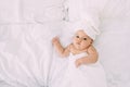 A portrait of a six-month-old girl lies on a white bed with a towel on her head and body. Hygiene and baby care concept.