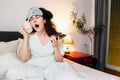 Portrait of sleeping yawning young woman wearing sleep mask call with cup and drink with phone while sitting in bed Royalty Free Stock Photo