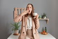 Portrait of sleepy brown haired woman wearing beige suit standing near table on kitchen at home, drinking coffee to have energy