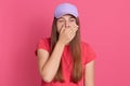 Portrait of sleepy long haired pretty teenager yawning, covering her mouth, having desire to sleep, wearing cap and t shirt,