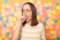 Portrait of sleepy overworked attractive woman wearing striped T-shirt standing against yellow wall covered with stickers, yawning Royalty Free Stock Photo