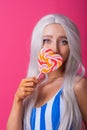 Portrait of a slender woman in a blonde wig posing with a lollipop on a pink background. Cute girl with blue eyes in a Royalty Free Stock Photo
