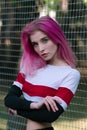 Portrait of slim fit girl cheerleader with pink hair and blue eyes in sportswear posing outdoors near fence at stadium