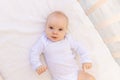 Portrait of a small baby girl 6 months old in a white bodysuit lying on her back on a white bed, space for text Royalty Free Stock Photo