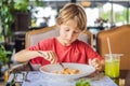 Portrait of small caucasian boy little child eating at restaurant in sunny day while sitting by the table alone - Royalty Free Stock Photo