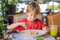 Portrait of small caucasian boy little child eating at restaurant in sunny day while sitting by the table alone - Royalty Free Stock Photo