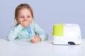 Portrait of small girl doing inhalation, sitting at table, wears in light blue turle neck sweater, has problem with healthy,