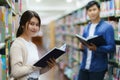 Portrait of Smart Asian man and woman university student reading book and looking at camera between bookshelves in campus library