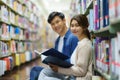 Portrait of Smart Asian man and woman university student reading book together and. looking at camera between bookshelves in Royalty Free Stock Photo