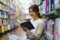 Portrait of Smart Asian woman university student reading book and looking at camera between bookshelves in campus library with Royalty Free Stock Photo