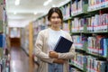 Portrait of Smart Asian woman university student reading book and looking at camera between bookshelves in campus library with Royalty Free Stock Photo