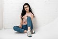 Portrait of smart beautiful brunette girl in eyeglasses with natural make-up sitting near white brick wall.