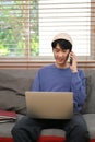 Smart Asian man on the phone with someone while using laptop on sofa in his living room Royalty Free Stock Photo