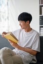 A happy young Asian man enjoys reading a book on a sofa in the living room