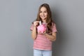 Little girl wearing striped T-shirt standing holding putting banknote into pink piggy bank in hands, Royalty Free Stock Photo