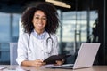 Portrait of a smiling African-American female doctor sitting in a white coat at the desk in the office, working on a Royalty Free Stock Photo