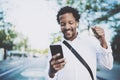 Portrait of Smiling African American man in headphones standidng in sunny street enjoying to music on his smart phone Royalty Free Stock Photo