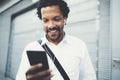 Portrait of Smiling African American man in headphones standidng in sunny street listening to songs on his mobile phone Royalty Free Stock Photo