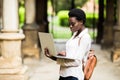 Portrait of smiling African american student doing homework outdoors using laptop for research. Positive dark skinned girl styling