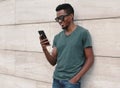 Portrait Smiling African Man With Phone Wearing T-shirt, Sunglasses On City Street Over Gray Wall