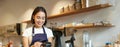 Portrait of smiling asian barista, coffee shop employee using POS terminal and credit card, helps client pay contactless