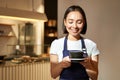 Portrait of smiling asian barista, girl in uniform, serving coffee, waitress holding cup of drink, working in cafe Royalty Free Stock Photo