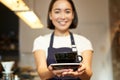 Portrait of smiling asian girl barista, waitress in cafe uniform, giving you cup of coffee, prepare drink for client Royalty Free Stock Photo