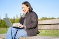 Portrait of smiling asian girl sits on bench in park, talks to friend online via laptop, video chats, using computer to Royalty Free Stock Photo