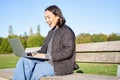 Portrait of smiling asian girl sits on bench in park, talks to friend online via laptop, video chats, using computer to Royalty Free Stock Photo