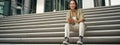 Portrait of smiling asian girl sits on stairs outdoors, sending message, using smartphone app, looking at mobile phone Royalty Free Stock Photo