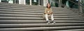 Portrait of smiling asian girl sits on stairs outdoors, sending message, using smartphone app, looking at mobile phone Royalty Free Stock Photo