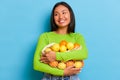 A portrait of smiling Asian woman holds a bunch of fresh oranges and lemons, looks excited, wears green cardigan Royalty Free Stock Photo