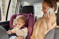 Portrait of smiling attractive mother using phone while sitting in car with her infant daughter on backseat, beautiful woman Royalty Free Stock Photo