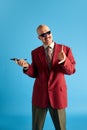 Portrait of smiling balded man with mustache dressed in red retro jacket with vintage mobile in hand over blue studio