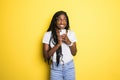 Portrait of a smiling beautiful african woman with afro hairstyle, holding mobile phone standing over yellow background Royalty Free Stock Photo